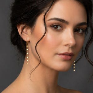 Può includere: Una donna con capelli scuri e pelle chiara indossa orecchini pendenti dorati. Gli orecchini presentano piccole perline rotonde. Indossa un abito verde scuro con spalle scoperte. Lo sfondo è grigio scuro uniforme.