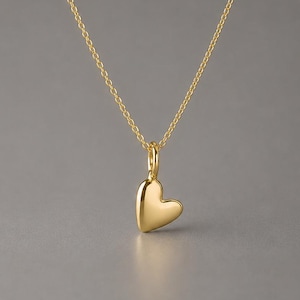 May include: A gold-toned necklace featuring a delicate chain and a small, smooth, solid heart-shaped pendant. The pendant hangs from a simple jump ring, showcasing a polished, reflective surface that catches the light, creating a minimalist look.