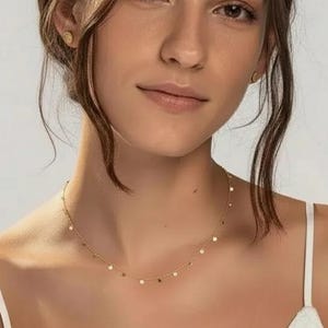 Peut inclure: Un collier en or délicat orné de petits charms ronds. La personne porte un haut blanc et a les cheveux et les yeux bruns. De petites boucles d'oreilles dorées sont également visibles. Le collier est près du cou.