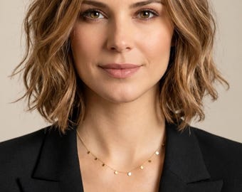 May include: A delicate gold chain necklace featuring small, evenly spaced circular gold discs. The necklace is worn around the neck, resting against the skin above a black blazer with a structured collar and lapels, set against a neutral background.
