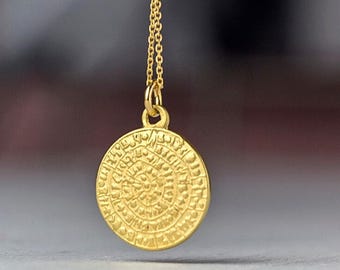 Pode incluir: Um colar com pingente dourado. O pingente é um disco circular com um design intrincado e texturizado. A corrente do colar também é dourada e delicada. O fundo está desfocado, destacando a joia.
