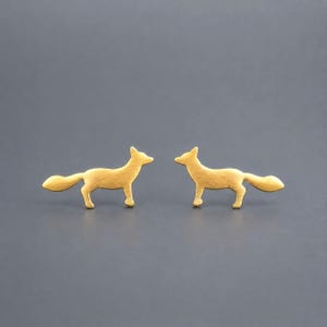 May include: A pair of gold-toned stud earrings shaped like foxes. Each earring features a stylized fox silhouette in a matte finish, showing the animal in a standing profile position with its tail extended horizontally behind its body.