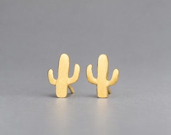 May include: A pair of gold-toned stud earrings shaped like saguaro cacti. Each earring features a flat, vertical central column with two smaller, curved arms extending from the sides, finished with a smooth, matte metallic surface texture.