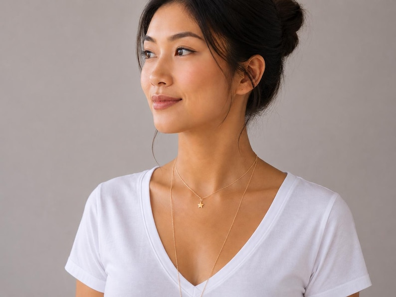 May include: A delicate gold-toned layered necklace featuring a small, five-pointed star charm on the shorter chain. The necklace consists of two thin, minimalist chains of varying lengths that rest against the collarbone and chest area.