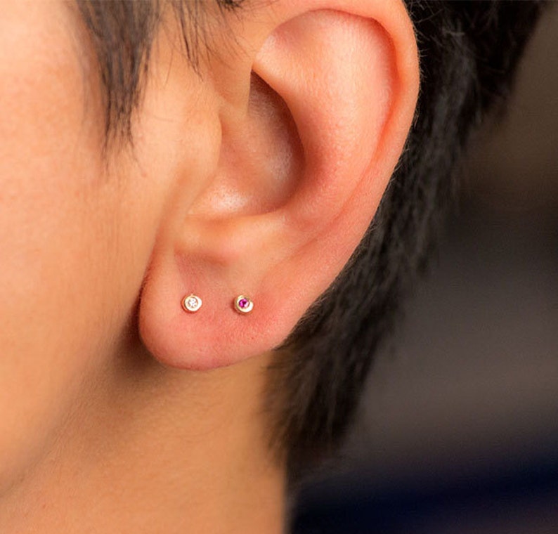 Może przedstawiać: Ucho z dwoma małymi kolczykami. Lewy kolczyk ma przezroczysty kamień, a prawy kolczyk ma r&oacute;żowy kamień.