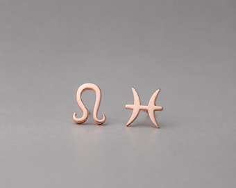 May include: A pair of rose gold stud earrings featuring the Leo zodiac symbol on the left and the Pisces zodiac symbol on the right. Each earring has a smooth, polished finish and a post backing, showcasing the distinct astrological glyphs.