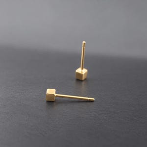 Può includere: Un paio di orecchini a lobo color oro, ciascuno caratterizzato da un piccolo ciondolo a forma di cubo liscio e lucido fissato a un perno dritto. Un orecchino è posizionato in verticale, mentre l'altro è disteso in orizzontale, mostrando la superficie metallica riflettente.