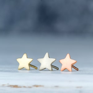 May include: Three star-shaped stud earrings arranged in a row. The first earring is gold-toned, the second is silver-toned, and the third is copper-toned. Each earring features a smooth, matte finish and a post backing for secure attachment.