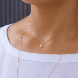 May include: A delicate gold-toned necklace featuring a small, five-pointed star charm suspended from a thin, fine chain. The necklace rests against the neck, showcasing a minimalist design with a polished metallic finish and a simple clasp link.