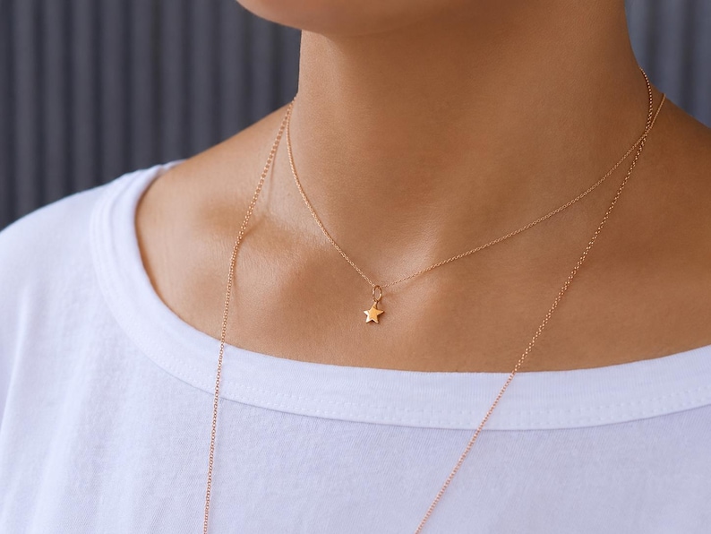 May include: A delicate gold-toned necklace featuring a small, five-pointed star charm suspended from a thin, fine chain. The necklace rests against the neck, showcasing a minimalist design with a polished metallic finish and a simple clasp link.
