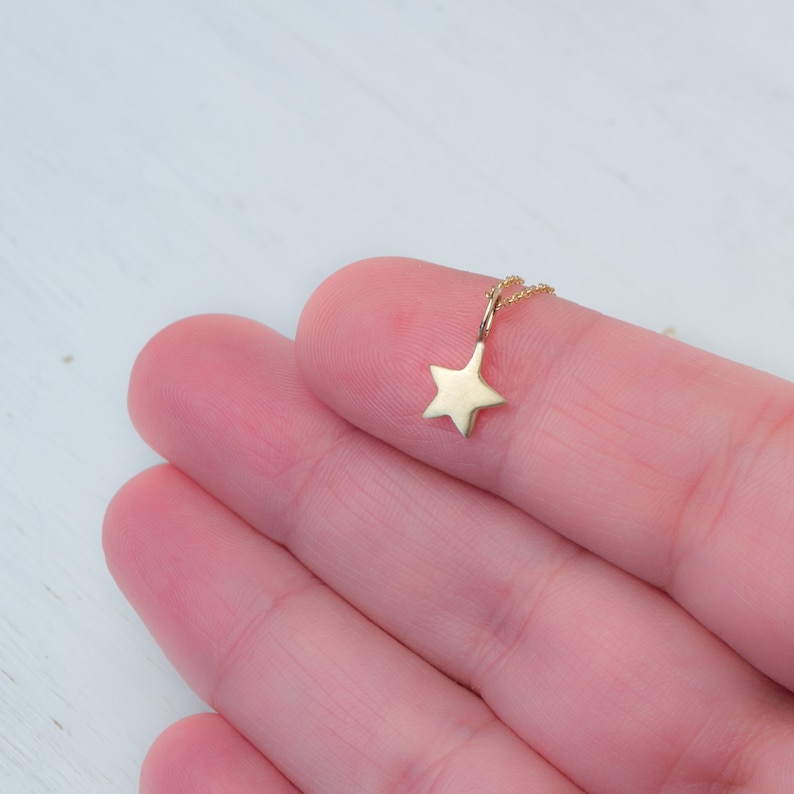 May include: A small gold star pendant attached to a delicate gold chain. The star has a smooth, polished finish and five points, suspended from a thin metal link that connects it to the fine chain links visible behind the pendant.