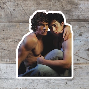 May include: A sticker featuring two men in a close embrace. One man is shirtless, with muscular arms and curly brown hair. The other man has dark hair and is wearing a white tank top and gray sweatpants.