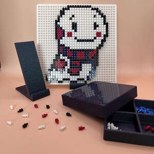 Könnte beinhalten: Eine Pixelkunst-Kreation eines Charakters mit weißem Gesicht, roten Wangen und einem Schal, dargestellt auf einer weißen Stecktafel. Ein schwarzer Telefonständer, eine dunkelblaue Box und verstreute Stifte in Rot, Weiß und Schwarz befinden sich im Vordergrund.
