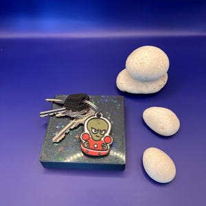 May include: A set of keys with a black Thule key fob and a cartoon alien keychain. The alien is green with a red outfit. The keys rest on a dark, glittery square. Three smooth, white stones are also present.