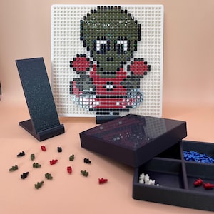 Op de afbeelding: Een pixel art ontwerp van een zombie op een wit pegboard. De zombie is groen, rood en zwart. Een zwarte telefoonstandaard, een donkerblauwe opbergdoos en diverse gekleurde pinnen staan op de voorgrond. De opbergdoos is open en toont compartimenten met pinnen.