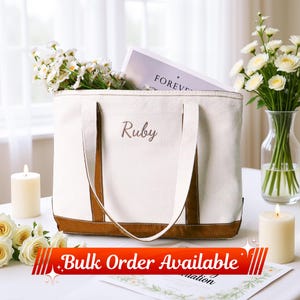 Puede incluir: Bolso tote color crema con detalles y asas marrones, bordado con el nombre "Ruby". La bolsa está llena de flores blancas y un libro con la palabra "FOREVER". La imagen también incluye velas y un arreglo floral.