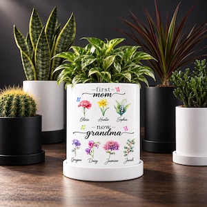 Könnte beinhalten: Ein weißer Keramik-Pflanztopf mit der Aufschrift first mom now grandma, verziert mit Aquarellillustrationen verschiedener Blumen und den Namen Olivia, Hunter, Sophia, Grayson, Daisy, Jasmine und Jonah sowie kleinen bunten Schmetterlingen.