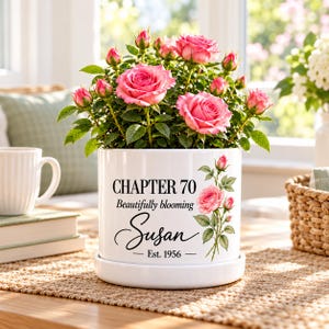 Könnte beinhalten: Ein weißer Keramikübertopf mit einem Miniatur-Rosenstrauch mit rosa Blüten. Der Übertopf ist mit dem Text "CHAPTER 70", "Beautifully blooming", "Susan" und "Est. 1956" sowie einer botanischen Illustration einer rosa Rose bedruckt.