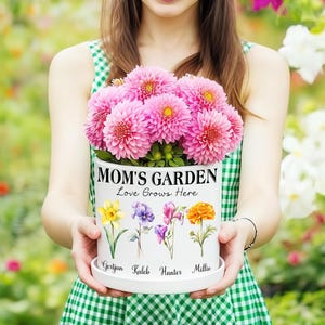 May include: A white ceramic flower pot with the text "MOM'S GARDEN Love Grows Here" and floral illustrations. The pot is filled with vibrant pink dahlias. The names Gertjan, Kaleb, Hunter, and Millie are printed below the flowers.