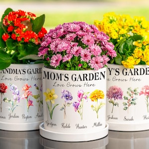 Könnte beinhalten: Drei weiße Keramik-Blumentöpfe mit bunten Blumen. Jeder Topf ist mit floralen Illustrationen und den Worten "GRANDMA'S GARDEN", "MOM'S GARDEN" und "Y'S GARDEN" verziert. Der Text "Love Grows Here" ist ebenfalls auf jedem Topf vorhanden.