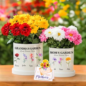 May include: Two white ceramic flower pots with the text "Grandma's Garden Love Grows Here" and "Mom's Garden Love Grows Here". Each pot has a row of flowers with names underneath. The pots are filled with colourful flowers. A card with the text "I Love You Happy Mother's Day" is in front of the pots.