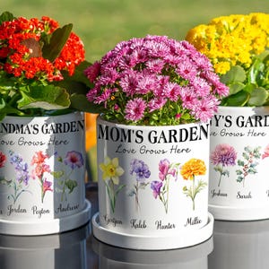 Op de afbeelding: Drie witte keramische bloempotten met kleurrijke bloemen. Elke pot is versierd met bloemenillustraties en de woorden "Grandma's Garden", "Mom's Garden" en "Amy's Garden". De potten hebben namen onderaan.