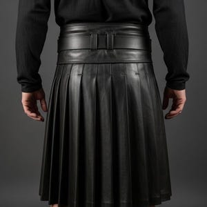 K&ouml;nnte beinhalten: Ein schwarzer Lederkilt mit tiefen Falten und einem breiten G&uuml;rtel. Der Kilt wird mit einem schwarzen Langarmshirt getragen. Der G&uuml;rtel hat eine rechteckige Schnalle. Der Kilt ist knielang.