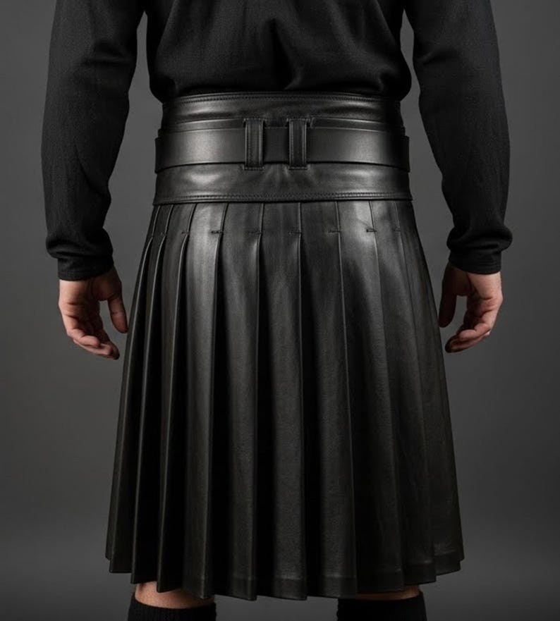 K&ouml;nnte beinhalten: Ein schwarzer Lederkilt mit tiefen Falten und einem breiten G&uuml;rtel. Der Kilt wird mit einem schwarzen Langarmshirt getragen. Der G&uuml;rtel hat eine rechteckige Schnalle. Der Kilt ist knielang.