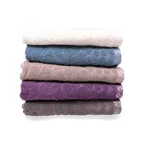 May include: A stack of six plush bath towels in various colors: white, blue, taupe, purple, and gray. Each towel features a textured pattern and a decorative floral border. The towels are neatly folded and stacked on top of each other.