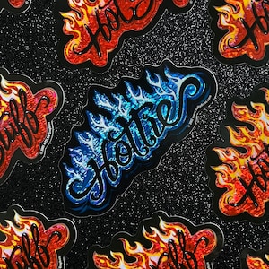 May include: A collection of stickers with the word "Hottie" in a flame design. The stickers are in red, orange, and blue, with a black outline. The background is black with glitter.