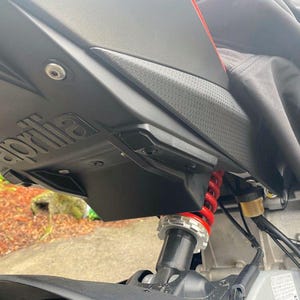 Puede incluir: Primer plano de la suspensión trasera y el chasis de una motocicleta negra. La motocicleta tiene un asiento negro y un amortiguador rojo. La palabra "Aprilia" es visible en el guardabarros trasero.