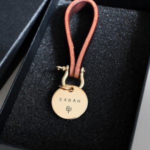 Puede incluir: Un llavero personalizado de color dorado con una correa de cuero, presentado en una caja de regalo negra. El llavero presenta el nombre "SARAH" y un símbolo de trébol de cuatro hojas. La correa de cuero es de color marrón cálido.
