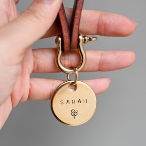 Könnte beinhalten: Ein goldfarbener, kreisförmiger Hundeanhänger mit dem Namen "SARAH" und einem vierblättrigen Kleeblattsymbol. Der Anhänger ist an einem braunen Lederriemen mit einem goldfarbenen Verschluss befestigt.