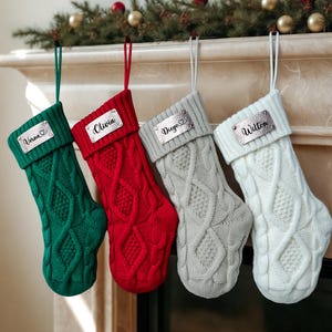 Puede incluir: Cuatro calcetines navideños de punto trenzado en verde, rojo, gris y blanco, cada uno con una placa con nombre plateada. Se muestran los nombres "Verna", "Olivia", "Diego" y "Wilton". Los calcetines cuelgan de una repisa decorada con vegetación y adornos.