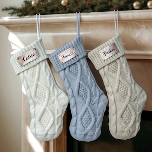 Könnte beinhalten: Drei strickverzierte Weihnachtsstrümpfe in Creme, Blau und Off-White hängen an einem Kaminsims. Jeder Strumpf hat ein metallisches Namensschild mit den Namen "Cedric", "Josue" und "Parker".