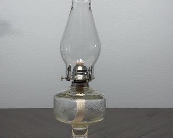 May include: A clear glass oil lamp featuring a textured, cylindrical base and a smooth, bulbous chimney. The lamp includes a silver-toned metal burner assembly with a visible white wick. The base has a decorative, embossed pattern around the bottom.