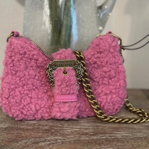 May include: A pink, fluffy shoulder bag with a gold chain strap and buckle closure. The bag features a textured, sherpa-like material and a rectangular shape. The buckle is engraved with "VERSACE JEANS COUTURE".