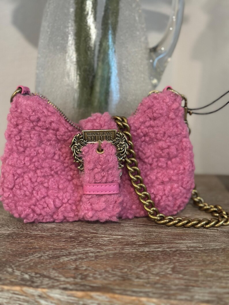 May include: A pink, fluffy shoulder bag with a gold chain strap and buckle closure. The bag features a textured, sherpa-like material and a rectangular shape. The buckle is engraved with "VERSACE JEANS COUTURE".