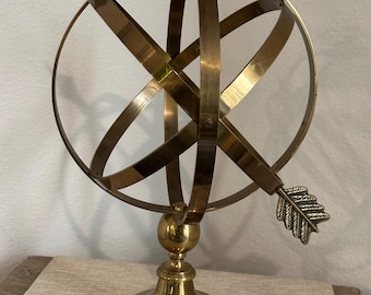 May include: A brass armillary sphere with a pointed arrow on a round base. The sphere is made of multiple intersecting rings.
