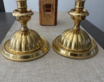 May include: Two ornate, gold-colored candle holders with a fluted base and a round, beaded edge. A decorative bottle with a dark stopper and a label with a crest is in the background. The candle holders are on a neutral-colored table runner.