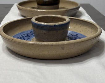 May include: Two handmade pottery serving dishes. The larger dish is tan with a blue interior and a central cup. The smaller dish is tan with a brown cup. Both dishes are on a white cloth.
