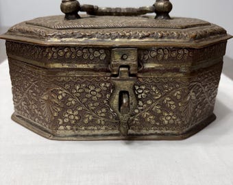 May include: An antique, octagonal metal box with intricate floral patterns and a hinged lid. The box features a handle and a clasp closure. The metal has a tarnished, aged appearance, suggesting it is vintage or an antique decorative item.