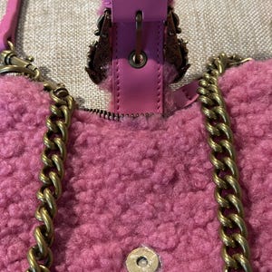 May include: Close-up of a pink, fluffy handbag with a gold chain strap. The bag features a magnetic clasp and decorative metal accents. The strap is a gold chain. The bag is made of a soft, textured material.