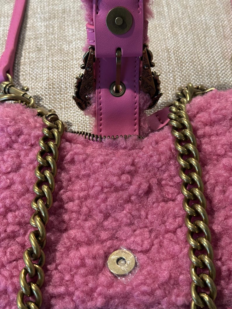May include: Close-up of a pink, fluffy handbag with a gold chain strap. The bag features a magnetic clasp and decorative metal accents. The strap is a gold chain. The bag is made of a soft, textured material.