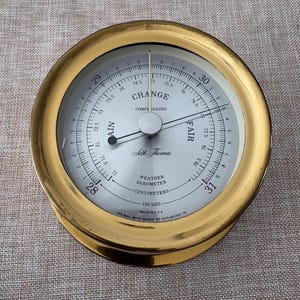 May include: A vintage Seth Thomas weather barometer with a gold-colored metal casing. The face displays measurements in inches and centimeters, with a needle pointing to the 'Fair' range. The text 'Change' and 'Compensated' are visible.
