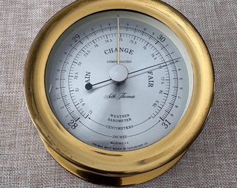 May include: A vintage Seth Thomas weather barometer with a gold-colored metal casing. The face displays measurements in inches and centimeters, with a needle pointing to the 'Fair' range. The text 'Change' and 'Compensated' are visible.