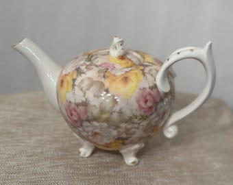 May include: A white porcelain teapot with a floral design featuring pink and yellow roses. The teapot has a round body with a curved spout and handle. It has three small feet and a lid with a knob.