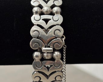 May include: A silver-toned bracelet with an ornate design. The bracelet features repeating openwork patterns with scroll and teardrop shapes, connected by spherical beads. A safety chain is attached.