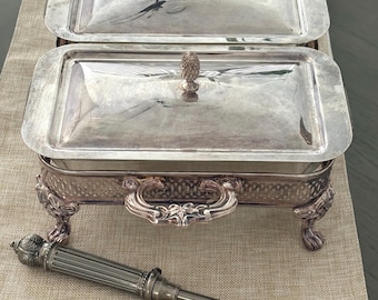 May include: A silver-plated rectangular chafing dish with two lids, each featuring a decorative finial handle. The base has ornate side handles and four claw-style feet. A long, slender silver-plated serving utensil with a fluted handle lies nearby.