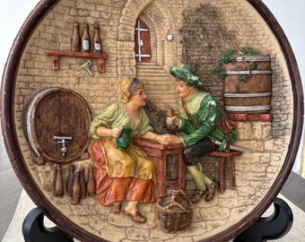 May include: A decorative, round wall plate with a relief scene of two people in a tavern setting. The plate features a brown border and a beige background with a brick wall design. The figures are dressed in period clothing, and there are barrels and bottles.